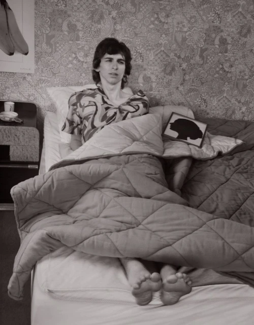 Monochrome photograph of a person in patterned shirt sitting in bed with a blanket, featuring floral wallpaper and a banana poster.