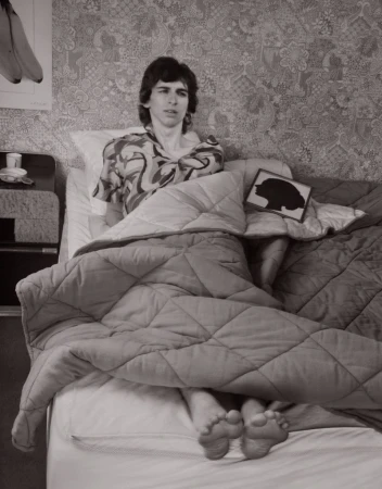 Monochrome photograph of a person in patterned shirt sitting in bed with a blanket, featuring floral wallpaper and a banana poster.