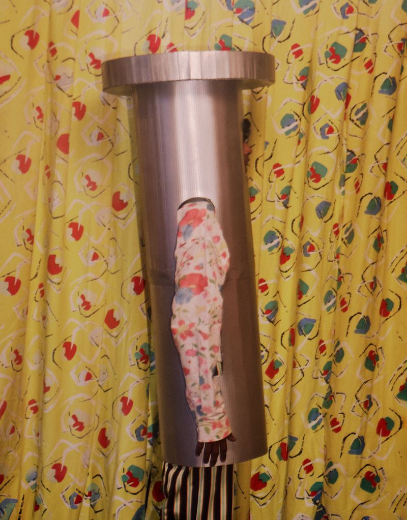 Abstract portrait of person wearing tube-shaped headwear, floral blouse, and vertically striped pants in front of patterned curtain.