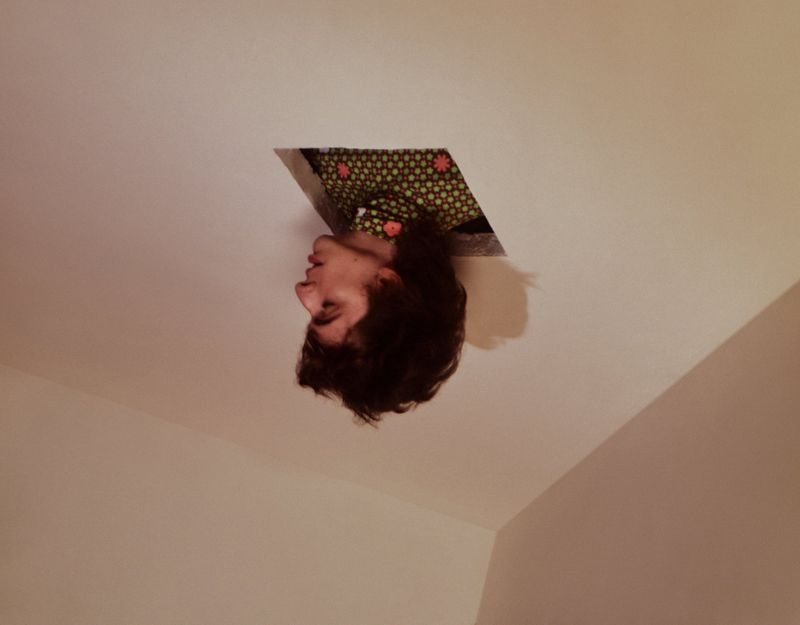 A person with curly dark hair looks out of a square ceiling opening, wearing a green patterned top, with creamy white walls.
