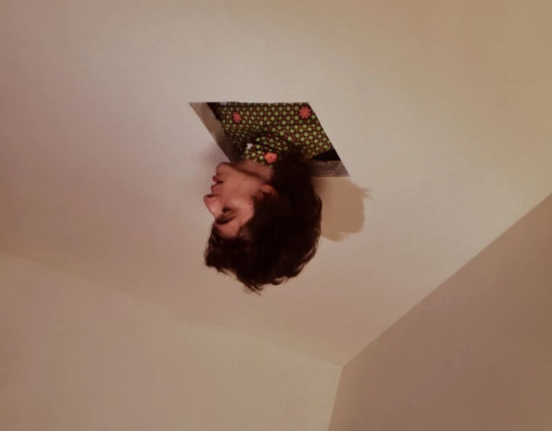 A person with curly dark hair looks out of a square ceiling opening, wearing a green patterned top, with creamy white walls.