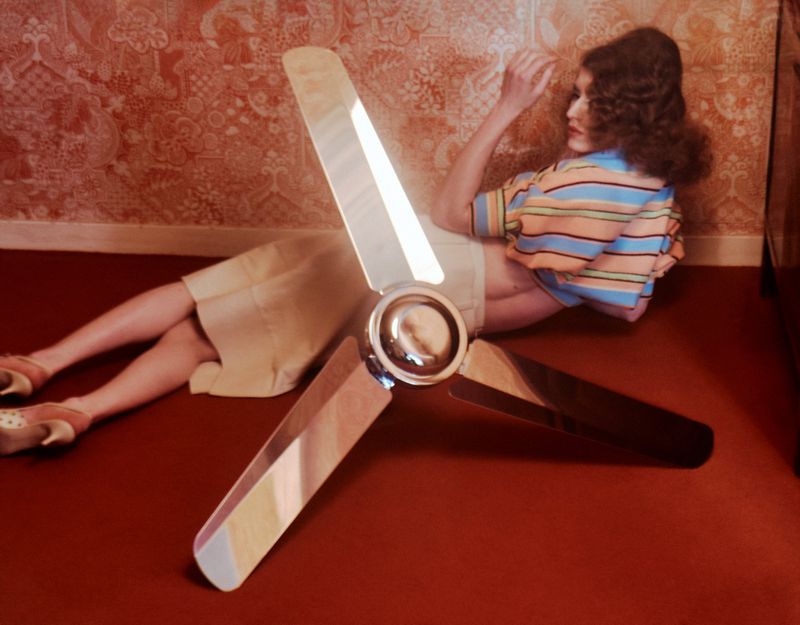 Retro indoor fashion shot of a woman reclining near a ceiling fan with warm lighting and vintage wallpaper, creating a nostalgic vibe.