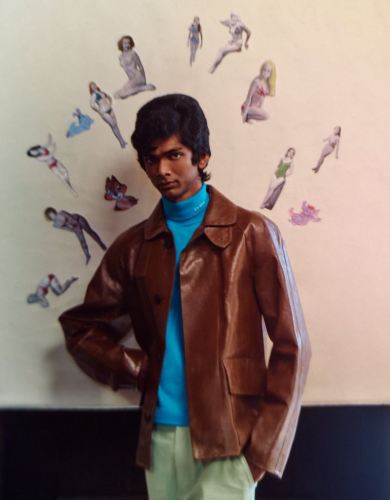 A young man with dark hair in a brown leather jacket and turquoise turtleneck posing in front of a wall with pin-up style art.