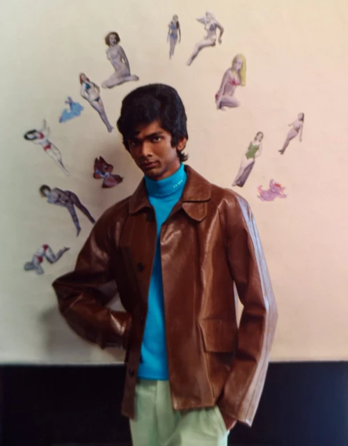 A young man with dark hair in a brown leather jacket and turquoise turtleneck posing in front of a wall with pin-up style art.