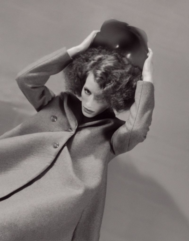 Black and white fashion shot of a slender woman in a wool coat and floppy hat, posed against a grey backdrop with soft, diffused lighting.