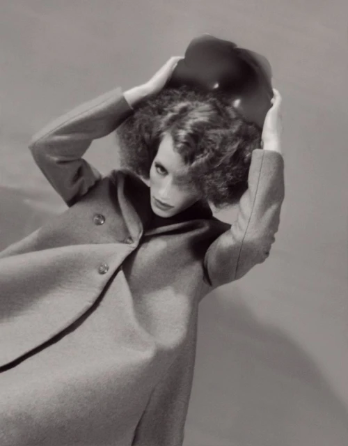 Black and white fashion shot of a slender woman in a wool coat and floppy hat, posed against a grey backdrop with soft, diffused lighting.