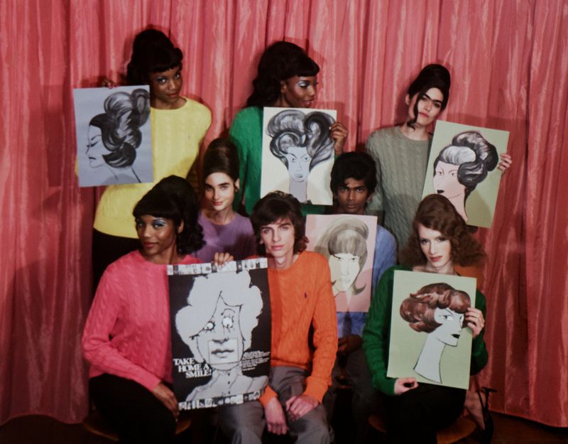 Group of young adult models in retro-style sweaters and updos holding art portraits in front of a pink curtain backdrop.