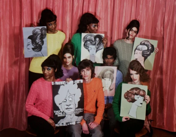 Group of young adult models in retro-style sweaters and updos holding art portraits in front of a pink curtain backdrop.