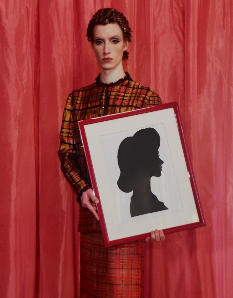 Adult poses in front of pink curtain holding a framed silhouette art piece. The model wears a vintage orange plaid skirt and jacket.