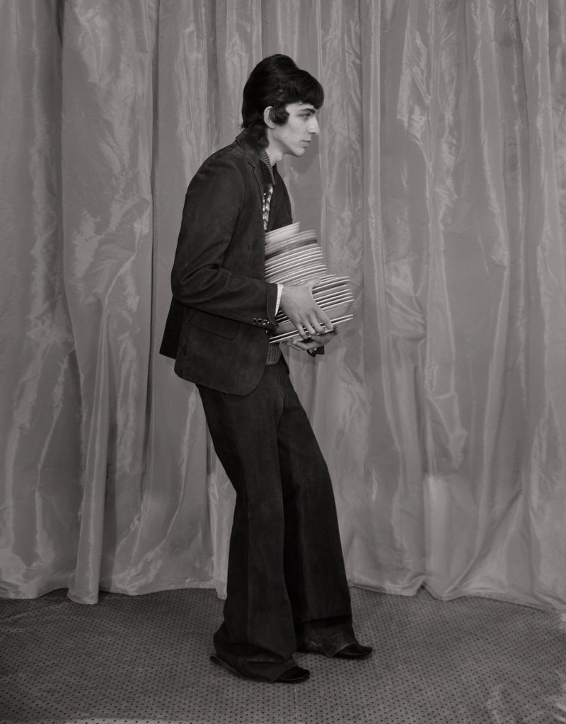 Monochromatic portrait of an adult male with dark hair carrying a stack of striped bowls in front of a draped backdrop.