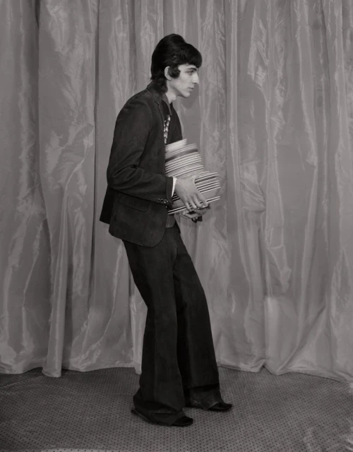Monochromatic portrait of an adult male with dark hair carrying a stack of striped bowls in front of a draped backdrop.