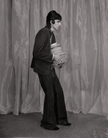 Monochromatic portrait of an adult male with dark hair carrying a stack of striped bowls in front of a draped backdrop.