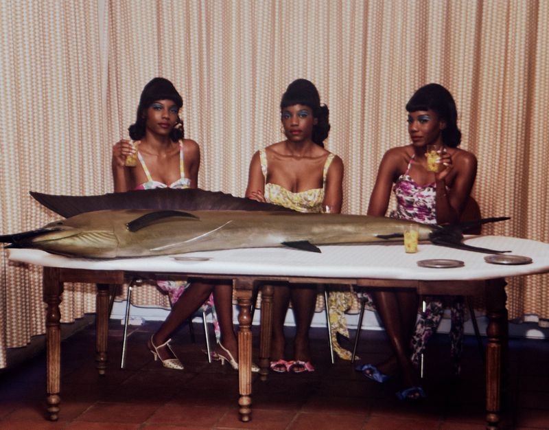 Three women in floral dresses sit at a table with a marlin. They're holding glasses, with plates and a patterned curtain in the background.