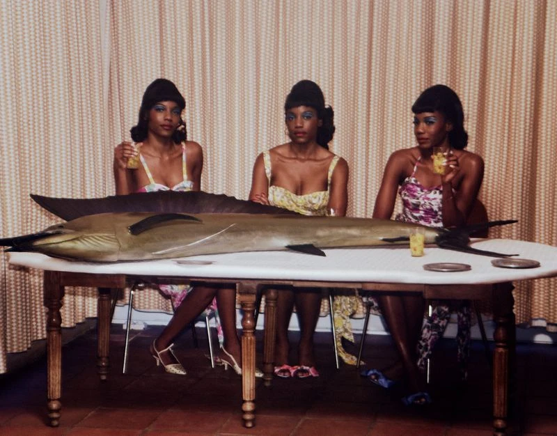 Three women in floral dresses sit at a table with a marlin. They're holding glasses, with plates and a patterned curtain in the background.