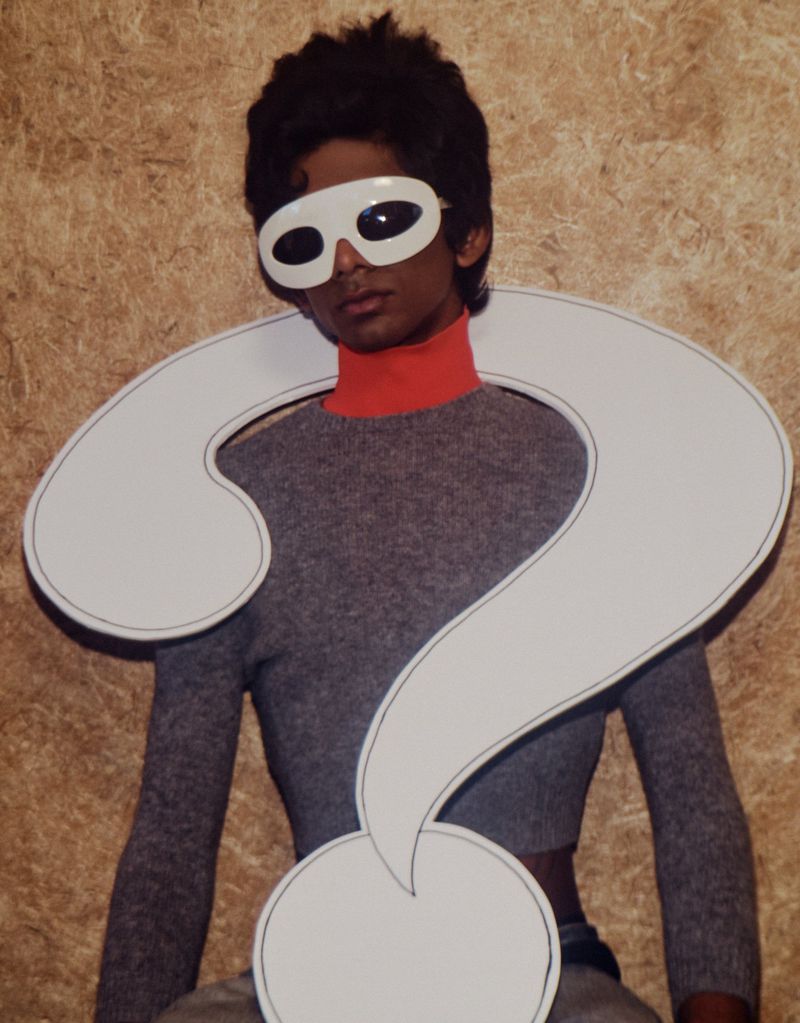 Person in a gray sweater and white glasses posing with a large white question mark in front of a mottled brown background.