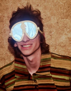A smiling young person wearing oversized white goggles and a striped multicolored collared shirt in front of a textured wall.