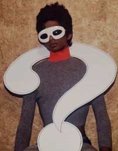 Model wearing a white eye mask and large question mark cutout prop over a cropped grey knit sweater against a chipboard wall.