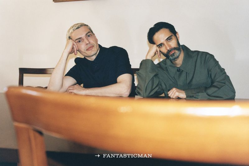 Two adult males sit at a wooden table in an indoor setting. The image features simple studio lighting and a minimalist background.