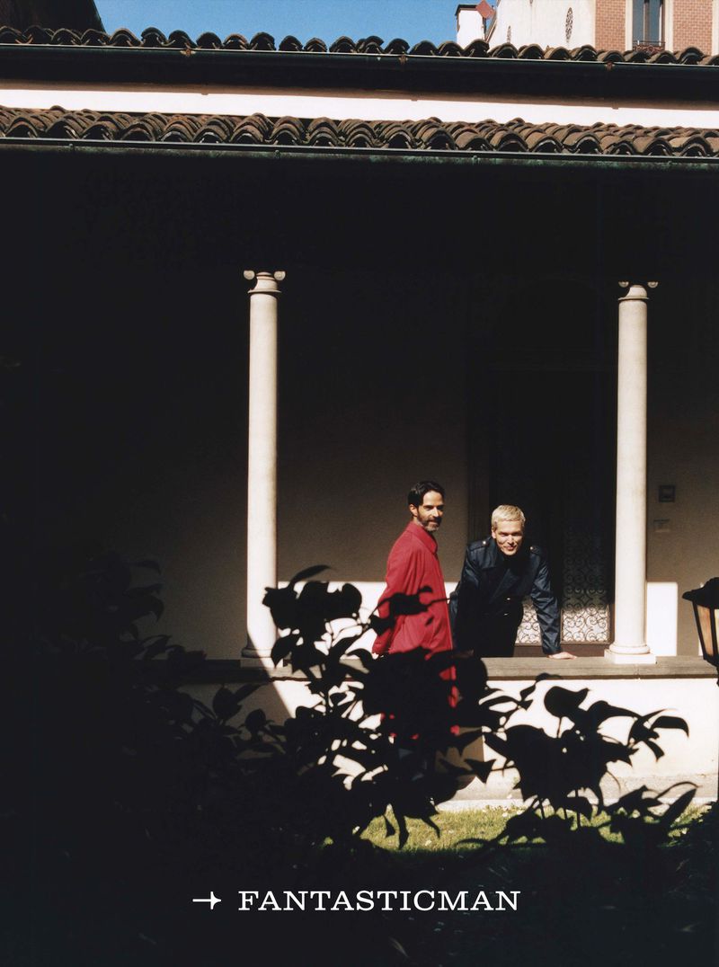 Two adult men pose in front of an arched opening with columns. One man wears a red shirt; the other wears a dark jacket. 