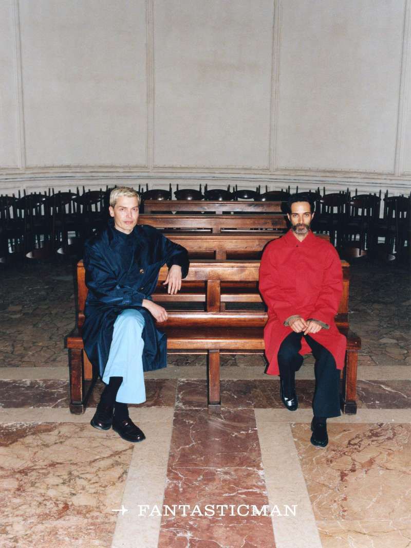 Two men seated on a wooden bench in a large hall, one in red and the other in blue, captured in a medium shot.