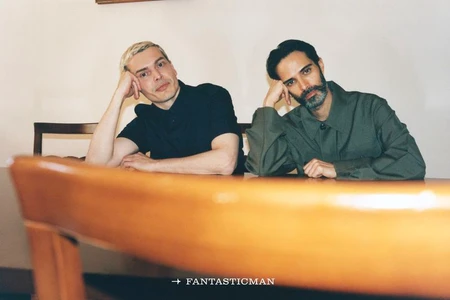 Two men seated at a wooden table leaning their heads on their hands indoors, with a blurred wooden chair in the foreground.