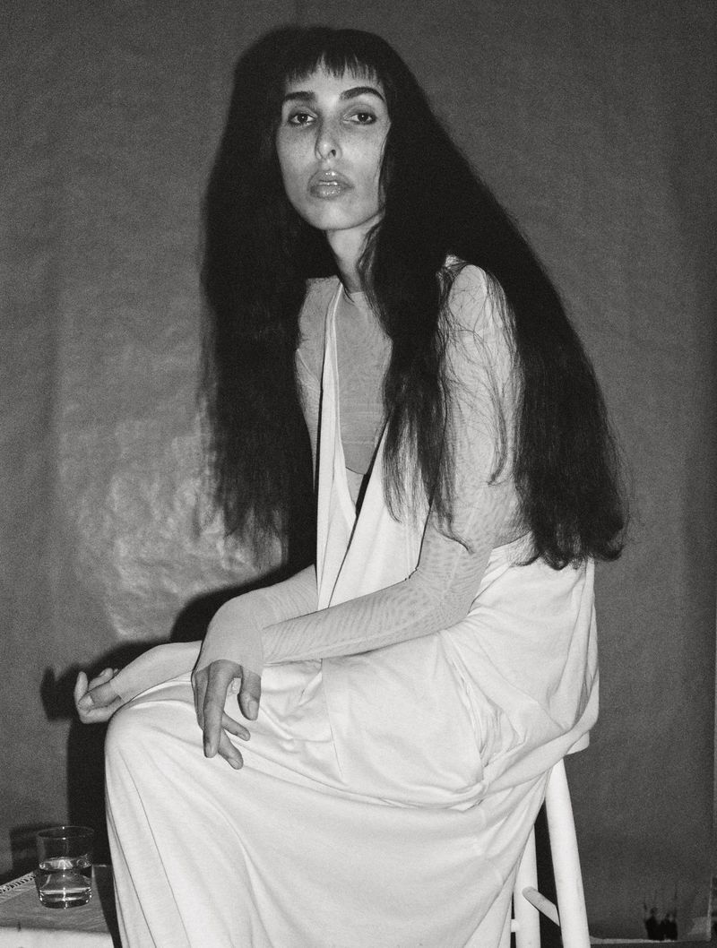 Model Raya Martigny seated on a white stool wearing a draped white dress in a black and white editorial portrait.