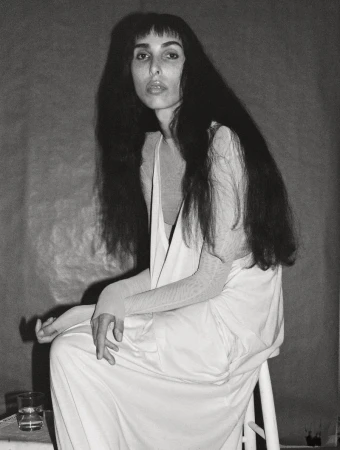 Model Raya Martigny seated on a white stool wearing a draped white dress in a black and white editorial portrait.