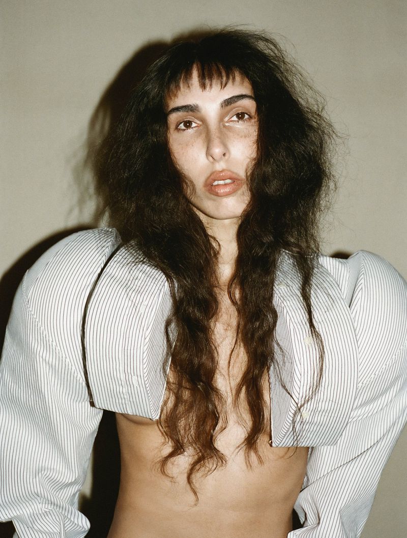 Woman with dark wavy hair and bangs in an unbuttoned striped shirt exposing her torso in a close-up studio portrait.
