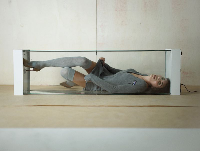 Woman laying in glass display case wearing gray cardigan and thigh-high socks with stiletto heels against neutral studio backdrop.