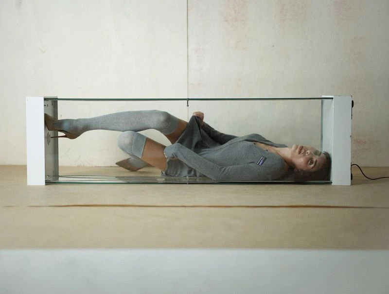 Woman laying in glass display case wearing gray cardigan and thigh-high socks with stiletto heels against neutral studio backdrop.