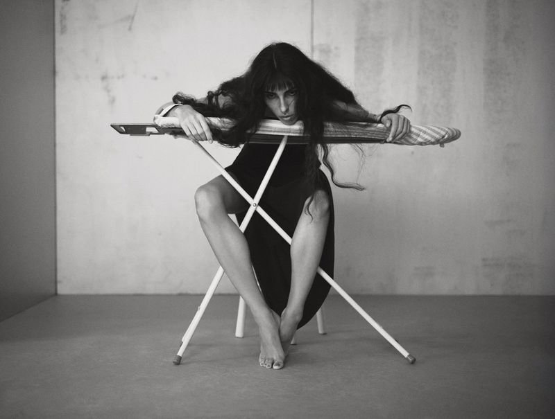 Black and white photograph of a woman with long dark hair reclining on an ironing board, looking at the camera with a neutral expression.