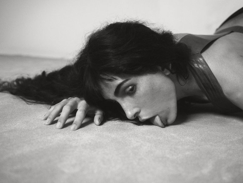 Black and white portrait of a woman lying down, her face resting on a textured surface, with dark hair flowing around her face and shoulders.