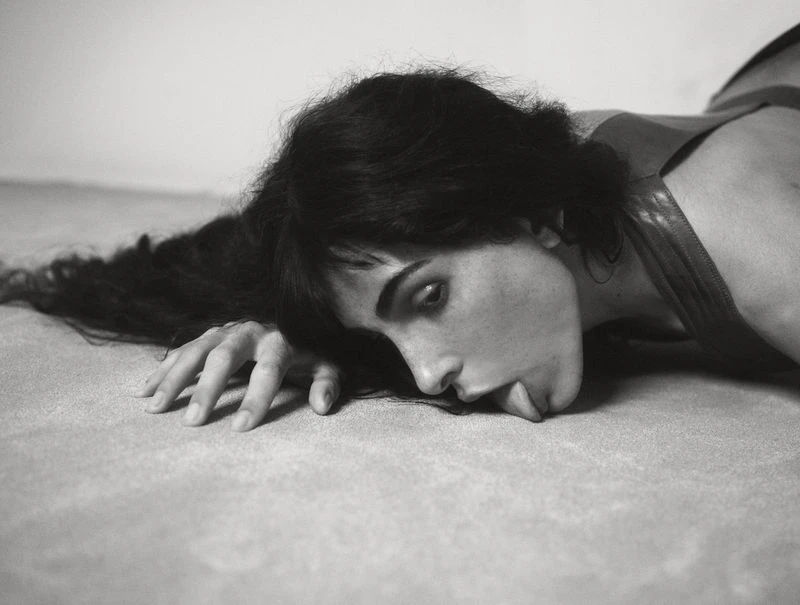 Black and white portrait of a woman lying down, her face resting on a textured surface, with dark hair flowing around her face and shoulders.