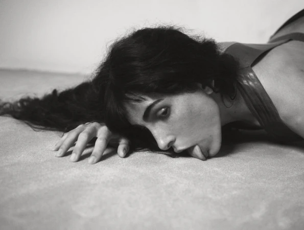 Black and white portrait of a woman lying down, her face resting on a textured surface, with dark hair flowing around her face and shoulders.