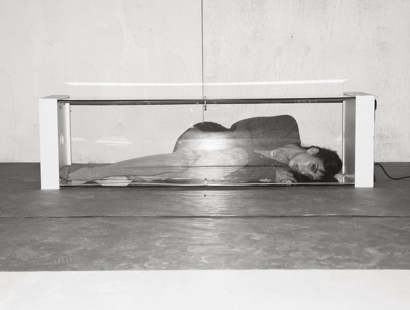 Monochrome image of a woman lying inside a transparent box-like structure with white edges, against a grey wall and floor.