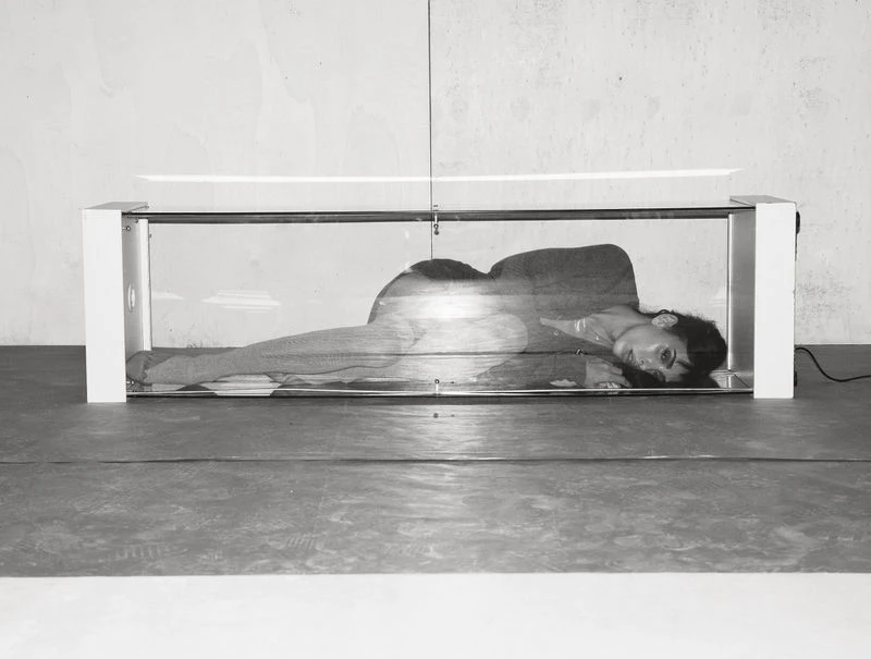 Monochrome image of a woman lying inside a transparent box-like structure with white edges, against a grey wall and floor.