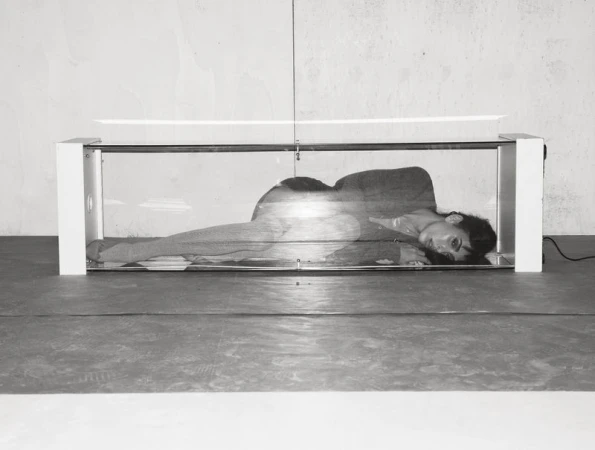 Monochrome image of a woman lying inside a transparent box-like structure with white edges, against a grey wall and floor.