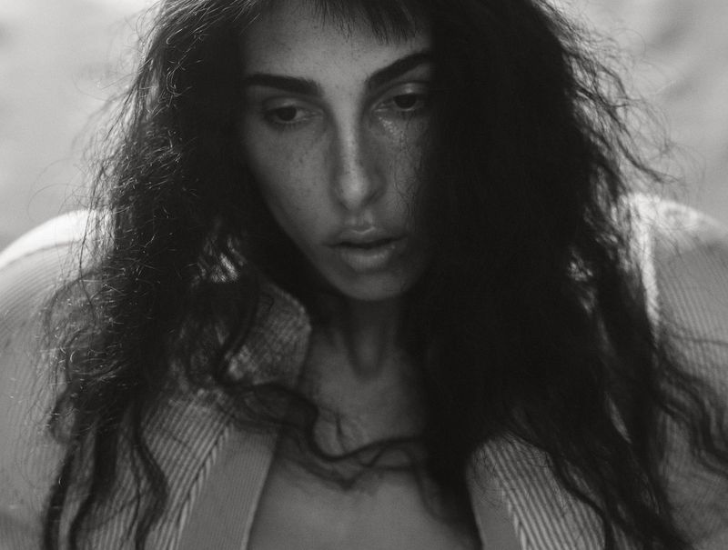 Portrait of a thoughtful adult female with long, wavy black hair and dark eyes, wearing a collared shirt, in a black and white aesthetic.