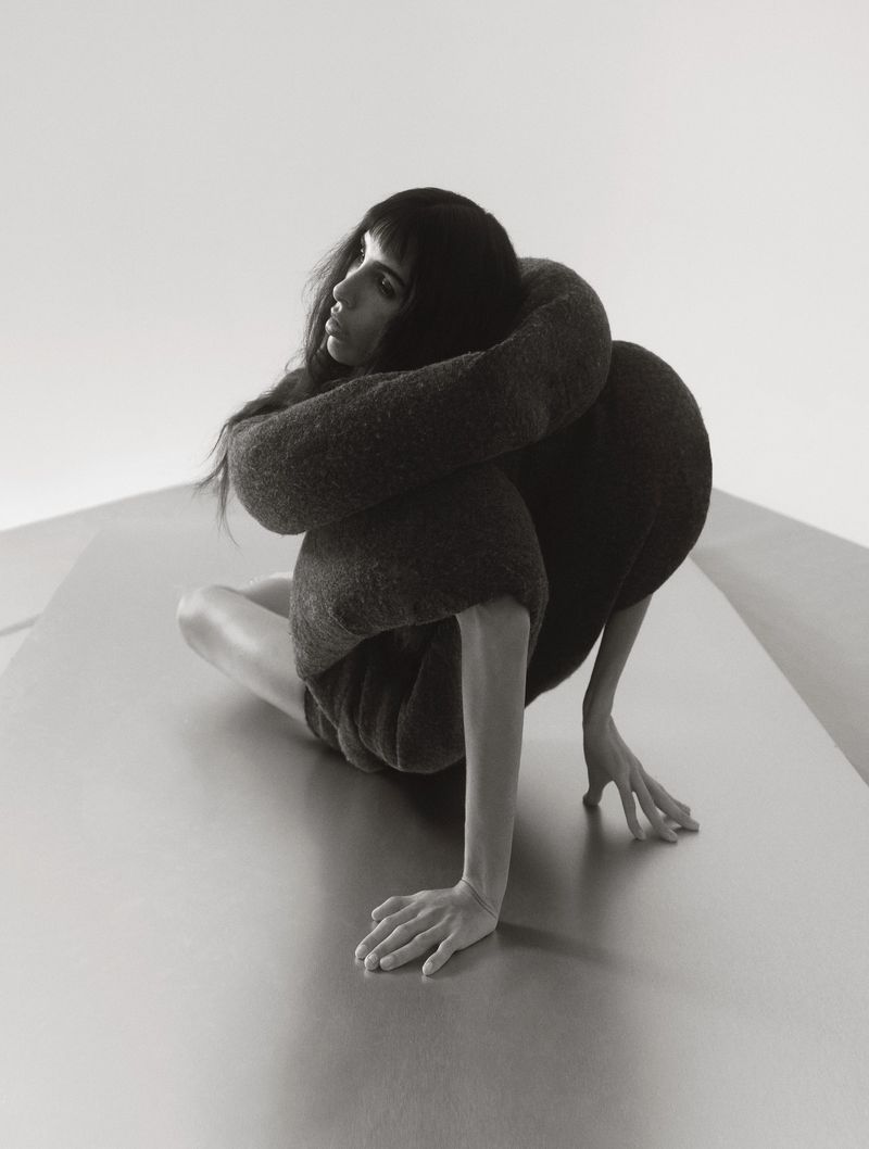 Monochromatic portrait of a woman sitting on a metallic surface, hugging herself, with a plain light grey background.
