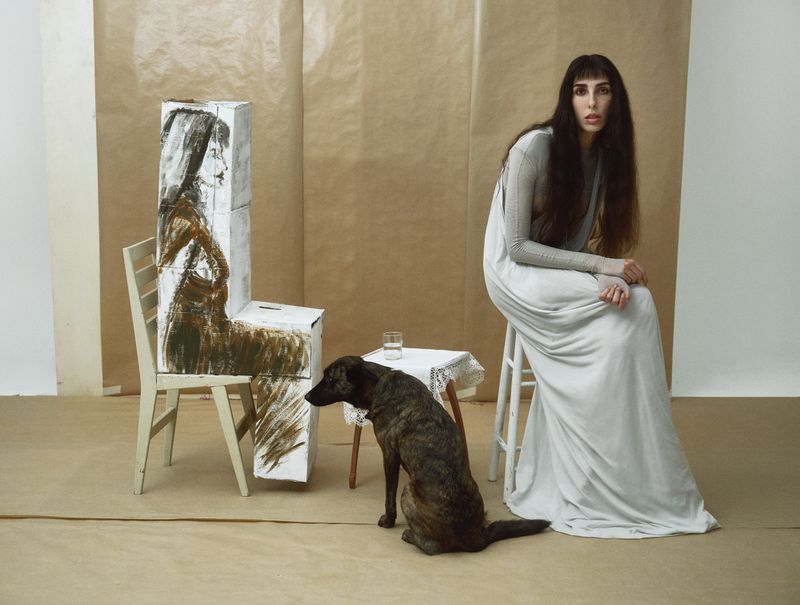 Indoor portrait of a woman in a white dress seated on a stool, accompanied by a dog and artwork on a chair, evoking a studio setting.