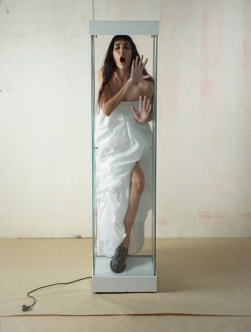 A woman in a white sheet stands inside a glass display case with her hands up against the glass. The scene is surreal and enigmatic.