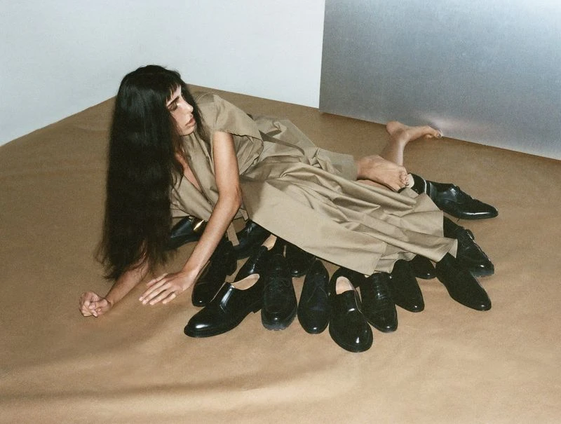 A woman with long dark hair in a khaki dress poses on the floor surrounded by an assortment of black dress shoes in a studio setting.