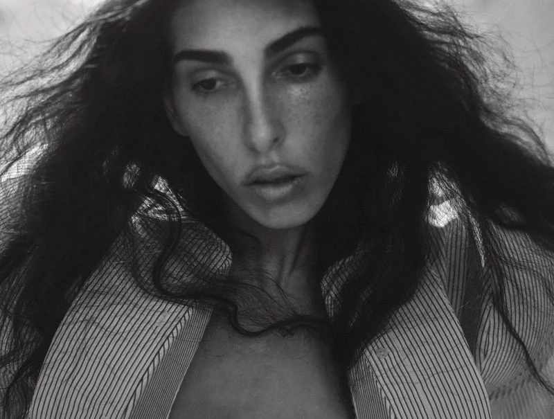 Striking black and white portrait of a woman with long dark hair wearing a striped shirt, captured in soft, natural light.