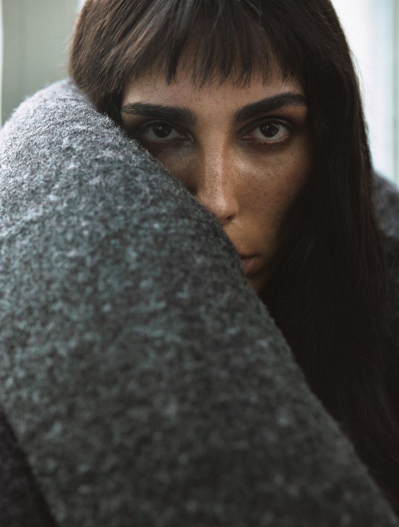 Portrait of an adult woman with freckles, dark hair, and bangs wrapped in a grey wool blanket looking directly at the camera indoors.
