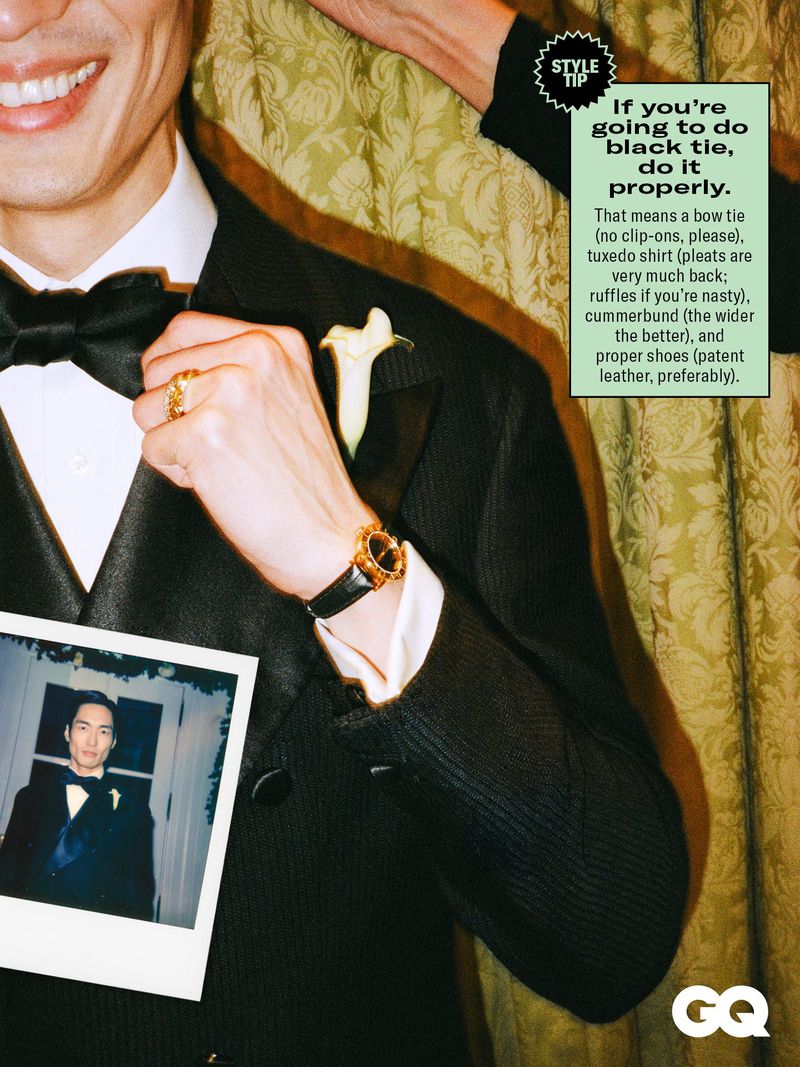Well-dressed man in a tuxedo with a bow tie and flower boutonniere, posing indoors with neutral patterned wall and a photograph.