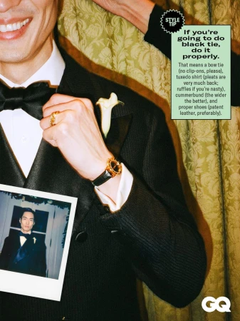 Model wearing a classic black tuxedo, bow tie, and gold watch, holding an instant photo against damask curtains for a GQ editorial.