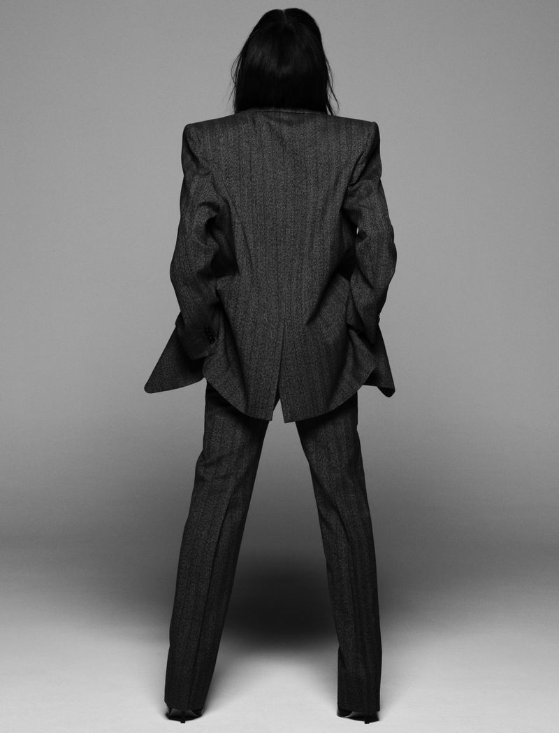 Back view of a person in a dark gray patterned suit with straight hair against a light gray seamless backdrop in a studio setting.