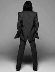 Full-length rear view of Victoria Beckham wearing an oversized dark grey tailored suit against a neutral studio backdrop.