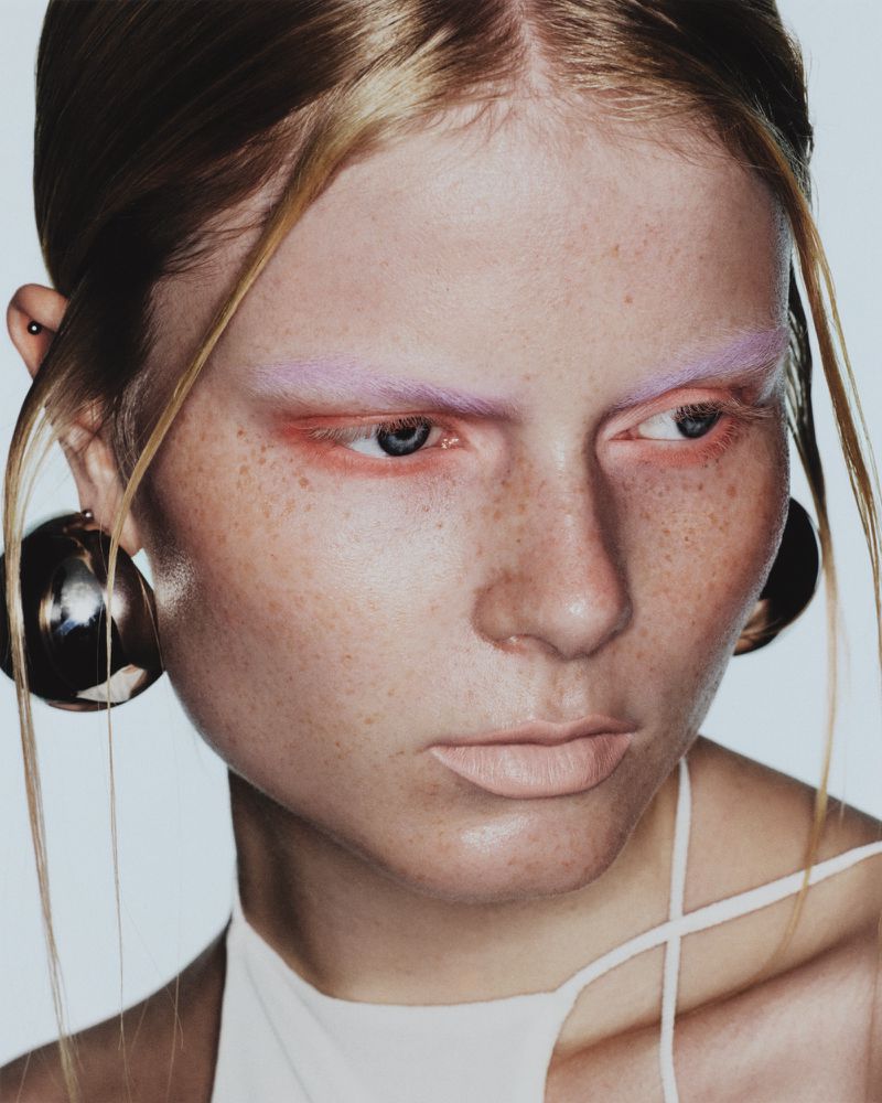 Close-up fashion portrait of a freckled, fair-skinned young woman with blonde hair and statement orb earrings, in a studio shot.