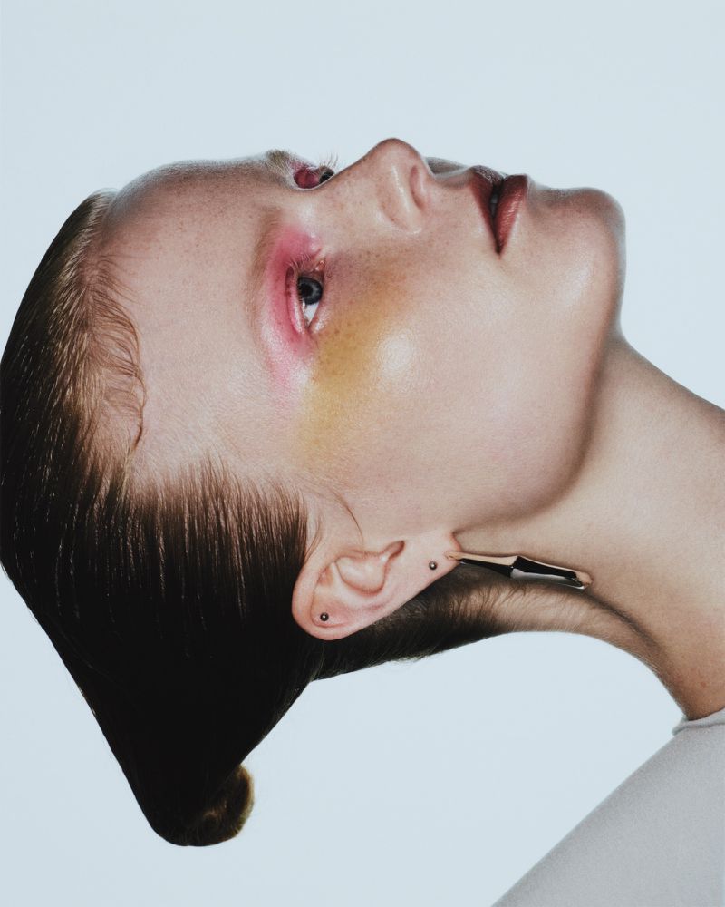 High-fashion portrait of a woman with sleek hair, side jewelry, and vibrant pink and gold makeup against a stark white background.